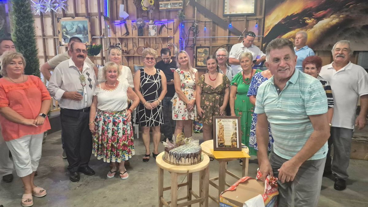 A large group gathered inside the decorated barn around a birthday cake, one woman in a floral Polish skirt and red coral beads.