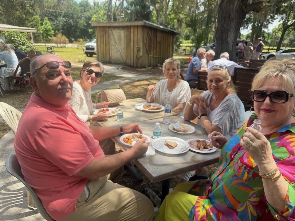 Guests of the club sitting at a table outdoors with plates of food, under oak trees.